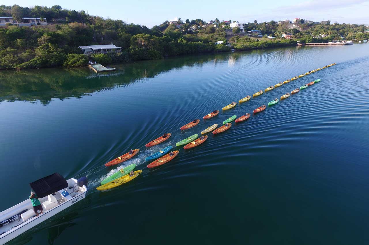 A motorboat pulls a long line of colorful kayaks, creating ripples in calm water. The kayaks are arranged in a straight line. Lush green hills and a few buildings are visible in the background under a clear sky.