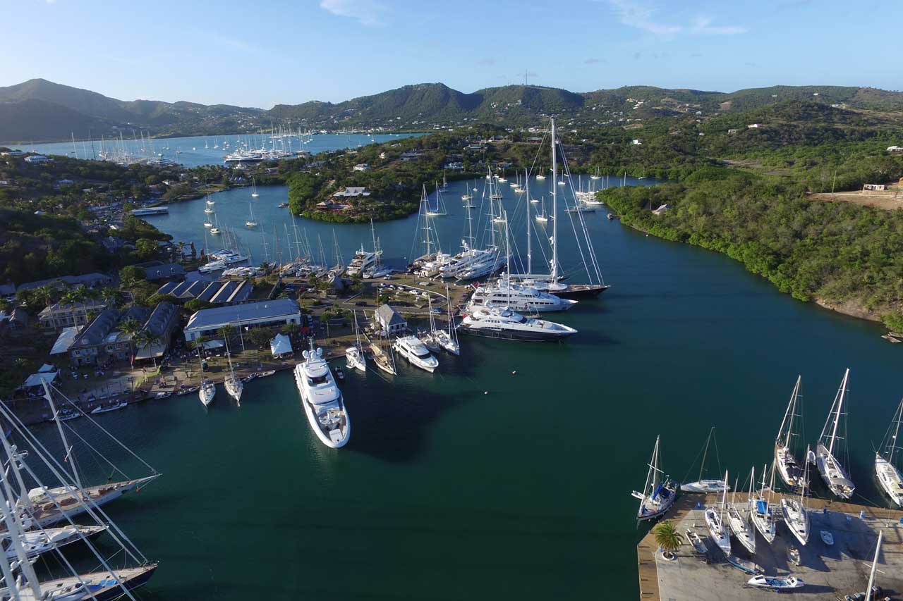 Aerial view of a harbor with numerous sailboats and yachts anchored. The water is surrounded by lush green hills and a small town. The image captures a tranquil scene with clear skies and serene waters.