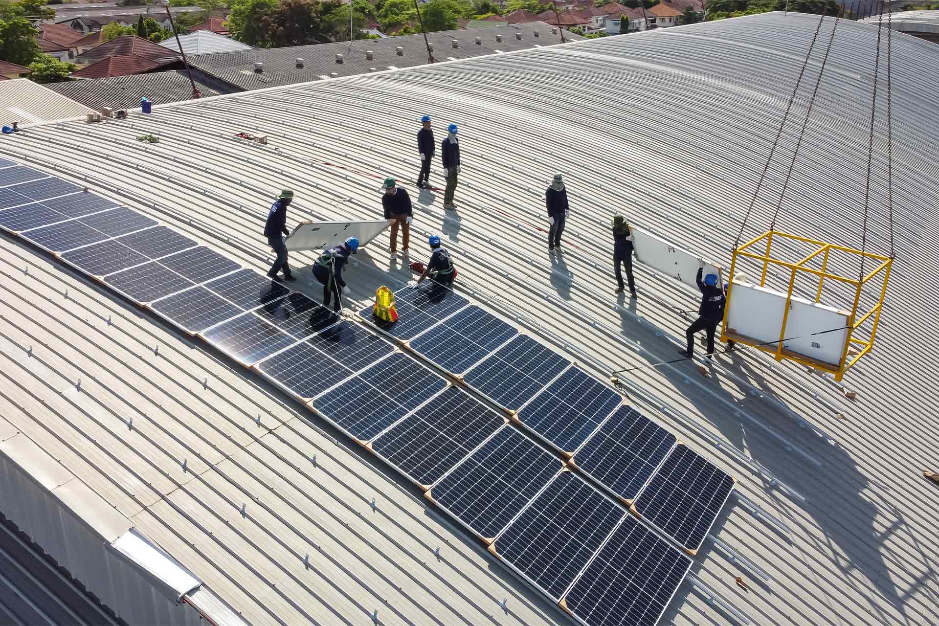 Workers wearing helmets install solar panels on a large corrugated metal rooftop, while a crane lifts additional panels in a yellow frame. Houses and trees are visible in the background.